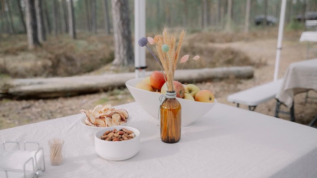 A rustic outdoor table setup featuring a bowl of apples, snacks, and a natural centerpiece amidst a forest backdrop.